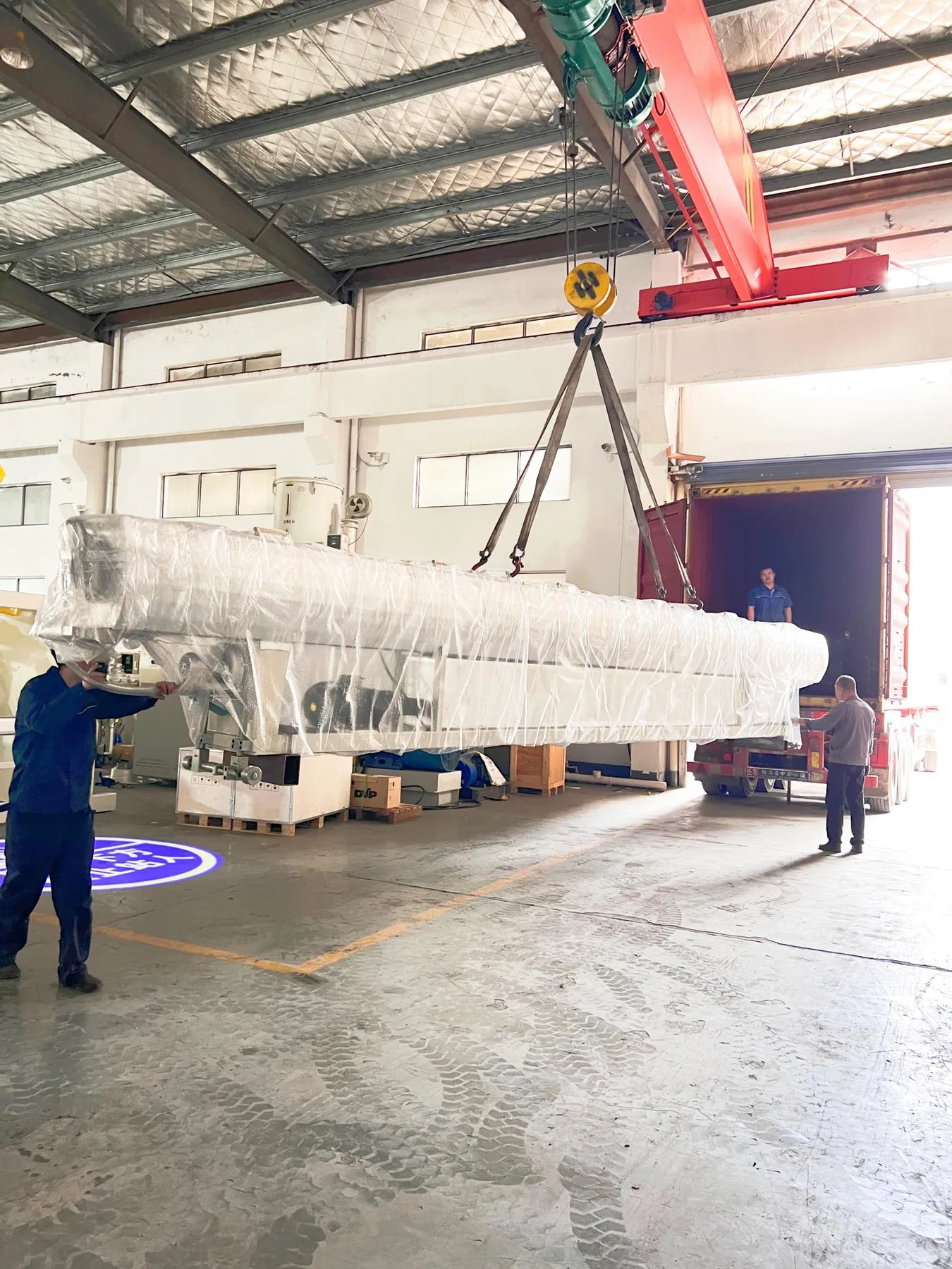 Overhead crane lifting a wrapped machine toward the container